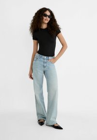 Schwarzes, eng anliegendes T-Shirt, hellblaue, hohe, weit geschnittene Jeans, schwarze, spitze Schuhe und schwarze Sonnenbrille vor einem weißen Hintergrund.