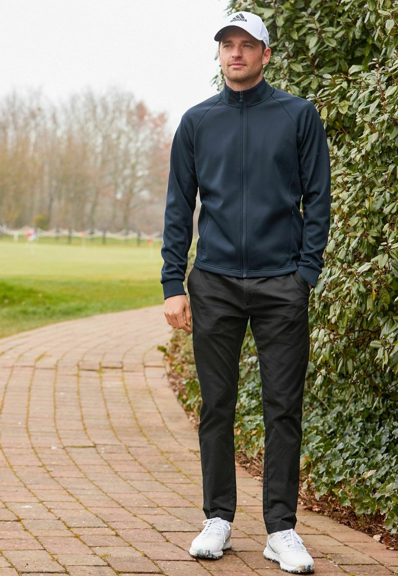 Chaqueta navy con cremallera, pantalones negros, zapatos blancos. Tela suave, corte atlético, con costuras sutiles. Gorra con logo que añade detalle.