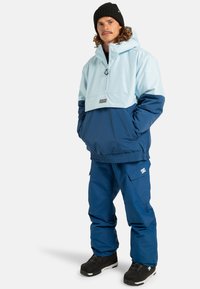 Jasnoniebieska i ciemnoniebieska kurtka z pół zamkiem i kieszeniami, w zestawieniu z dopasowanymi niebieskimi spodniami. Czarne buty i czarna czapka dopełniają stylizację.