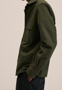 Camicia button-up verde oliva con maniche lunghe, tasca sul petto e bottoni neri. Tessuto liscio con una vestibilità strutturata.