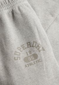 Gray sweatpants with a textured finish, featuring a soft elastic waistband, and a printed logo that reads "Superdry 1986 Athletic."