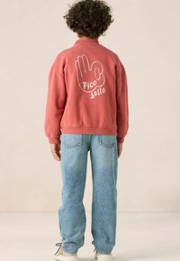 Sweatshirt corail avec un graphique de main blanche et le texte "pico bello" au dos. Associé à un jean bleu clair et des baskets blanches.