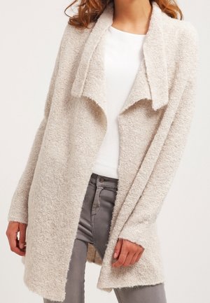 Femme portant un cardigan beige clair texturé à ouverture devant, sur une chemise blanche et un pantalon gris, avec des manches longues décontractées.