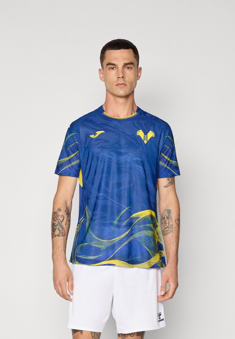 Maglietta sportiva blu a maniche corte con accenti gialli e un motivo a onda. Presenta un collo rotondo e un logo sul petto, abbinata a pantaloni corti bianchi.