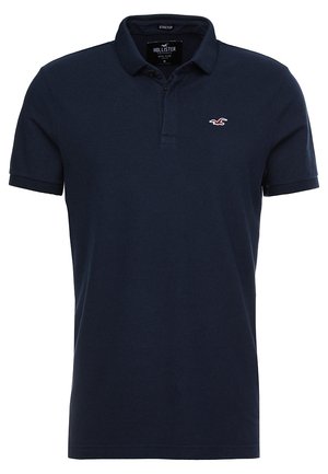 Polo de color azul marino hecho de tejido elástico. Presenta un cuello con solapa, mangas cortas y un pequeño logo rojo en el pecho.