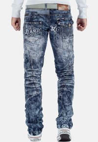 Jeans in denim blu sbiadito con cuciture intricate, molteplici tasche e dettagli decorativi sul retro. Vestibilità su misura con una leggera affusolatura.