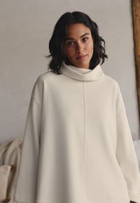Felpa a coste color crema con collo a imbuto, vestibilità oversize e spalle scese. Texture liscia e dettagli di cuciture minimi.