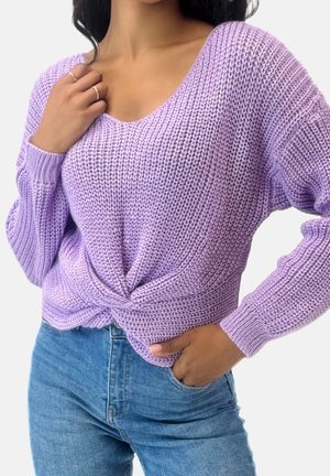 Mujer con un suéter de punto color lavanda con un detalle de torsión frontal y vaqueros azules, una mano en el bolsillo y la otra cerca del cuello.