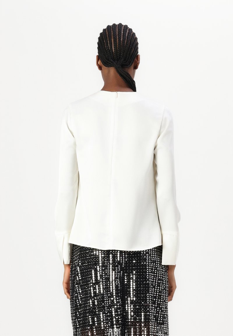 Biała bluzka z długim rękawem, okrągłym dekoltem i suwakiem z tyłu. Posiada gładką teksturę i lekko rozszerzany dół. W zestawie z czarną spódnicą pokrytą cekinami.