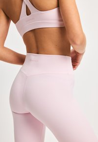 Lys rosa sportbh og høyhøydde leggings i et glatt, elastisk stoff; har merkevarenavn på kanten; minimal design uten mønstre.