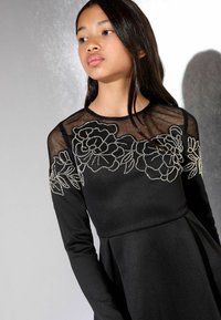 Fekete, hosszú ujjú ruha, átlátszó nyakkal és ezüsthímzéses virágmintával. Bővülő szoknya, puha textúrával.