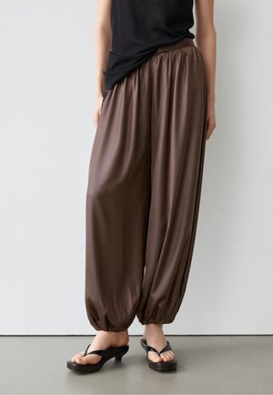 Personne portant un pantalon marron ample avec un ourlet froncé, des sandales à talons noires et un top noir sans manches devant un mur uni.