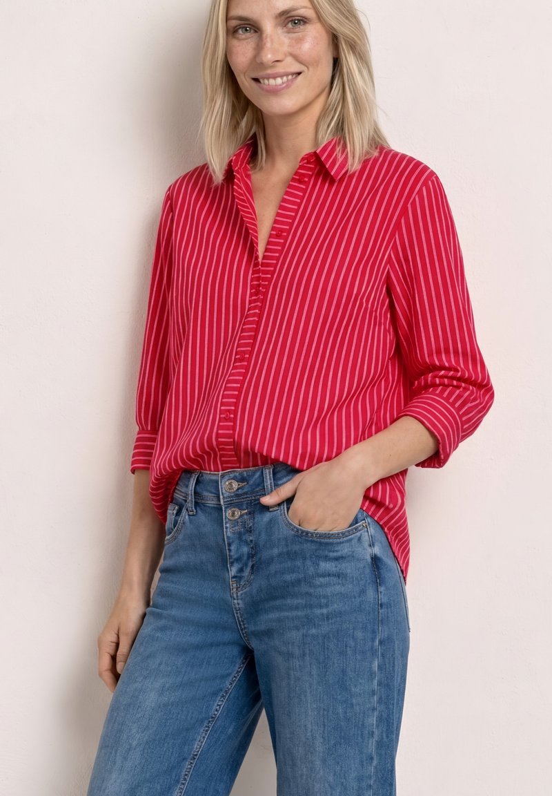 Femme portant une chemise à rayures rouges et blanches boutonnée avec les manches retroussées et un jean bleu taille haute, souriant avec une main dans la poche.