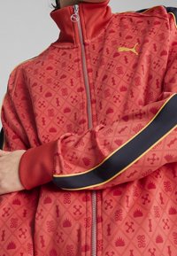 Chaqueta roja con cremallera y patrones geométricos, y cuello acanalado. Rayas negras y amarillas adornan las mangas. Tejido suave y texturizado.