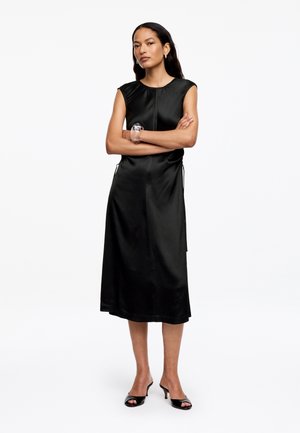 Kobieta stojąca z założonymi skrzyżowanymi ramionami, ubrana w czarną, bezrękawnikową sukienkę midi, czarne sandały na obcasie oraz przezroczystą bransoletkę na nadgarstku.