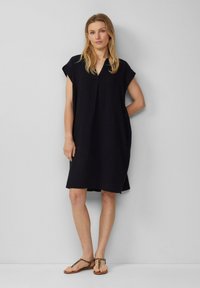 Schwarzes leichtes Kleid mit V-Ausschnitt und kurzen Ärmeln, das einen lockeren Schnitt und strukturierte Stoffe aufweist. Kombiniert mit flachen Sandalen.