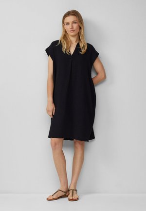 Schwarzes leichtes Kleid mit V-Ausschnitt und kurzen Ärmeln, das einen lockeren Schnitt und strukturierte Stoffe aufweist. Kombiniert mit flachen Sandalen.