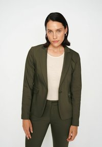 Olīvu zaļš pielāgots žakets ar noapaļotām apkaklēm un divām priekšējām kabatām, kombinēts ar vieglu bēšu pieguļošu blūzi. Gluda tekstūra, strukturēta piegriezuma.