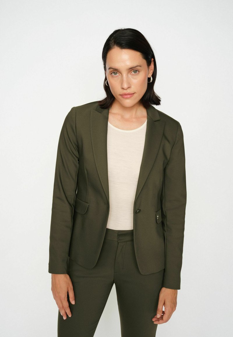 Olīvu zaļš pielāgots žakets ar noapaļotām apkaklēm un divām priekšējām kabatām, kombinēts ar vieglu bēšu pieguļošu blūzi. Gluda tekstūra, strukturēta piegriezuma.