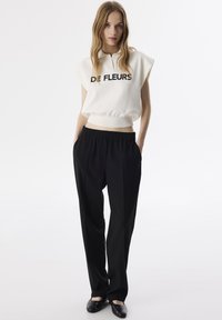 Crème mouwloos topje met "DE FLEURS" opdruk, gecombineerd met zwarte elastische taillebroek. Model draagt zwarte platte schoenen en staat tegen een effen achtergrond.