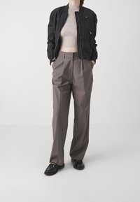 Černá bomber bunda se zipem, béžový žebrovaný crop top, šedé skládací kalhoty a černé mokasíny se zlatými akcenty.