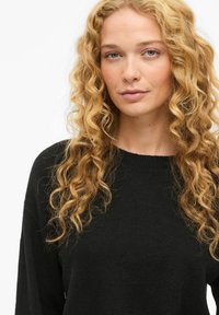 Kvinna med långt, lockigt blont hår som bär en svart tröja och ser något åt sidan med ett neutralt uttryck mot en vit bakgrund.