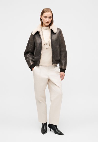 Καφέ δερμάτινο μπουφάν τύπου bomber με μπεζ παλτό shearling στο γιακά, φορεμένο πάνω από ένα ανοιχτόχρωμο πουλόβερ και εκρού παντελόνι, σε συνδυασμό με μαύρες μυτερές γόβες.