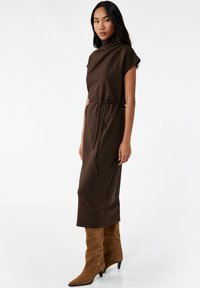 Vestido marrón de longitud hasta la rodilla con un cuello alto, mangas cortas y cintura con cordón. Llevado con botas altas de ante en color beige. Diseño sencillo y ajustado.