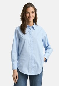 Light blue button-up shirt with vertical white stripes, long sleeves, rounded hem, and subtle detailing on fabric; paired with blue jeans.