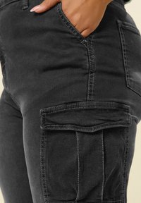 Mustat farkkuhousut, joissa on sivutasku, kontrastiväriompeleet ja sileä pinta. Lisätaskuyksityiskohta näkyvissä.