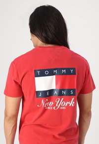 Rød bomulls T-skjorte med grafisk trykk på baksiden med marineblå og hvite rektangler, tekst "TOMMY JEANS New York siden 1985."