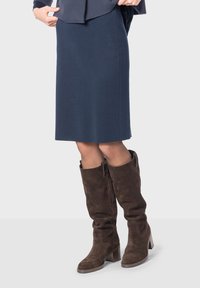 Jupe droite bleu marine à hauteur du genou associée �à des bottes en suède marron hautes avec un talon carré et un zip latéral. Texture lisse, design simple.