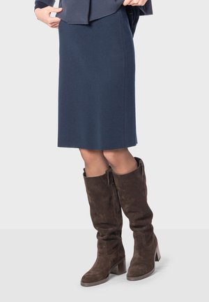Jupe droite bleu marine à hauteur du genou associée à des bottes en suède marron hautes avec un talon carré et un zip latéral. Texture lisse, design simple.
