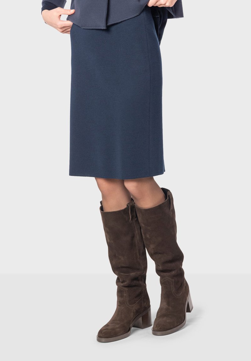 Jupe droite bleu marine à hauteur du genou associée à des bottes en suède marron hautes avec un talon carré et un zip latéral. Texture lisse, design simple.