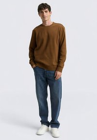 Felpa marrone con collo rotondo, tessuto strutturato, vestibilità ampia, abbinata a jeans blu e sneakers bianche, in piedi contro uno sfondo neutro.