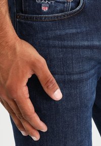 Los pantalones vaqueros de mezclilla azul oscuro presentan un corte slim, un bolsillo frontal y un pequeño logo de GANT en la cintura con costuras detalladas. Mano apoyada en el muslo.