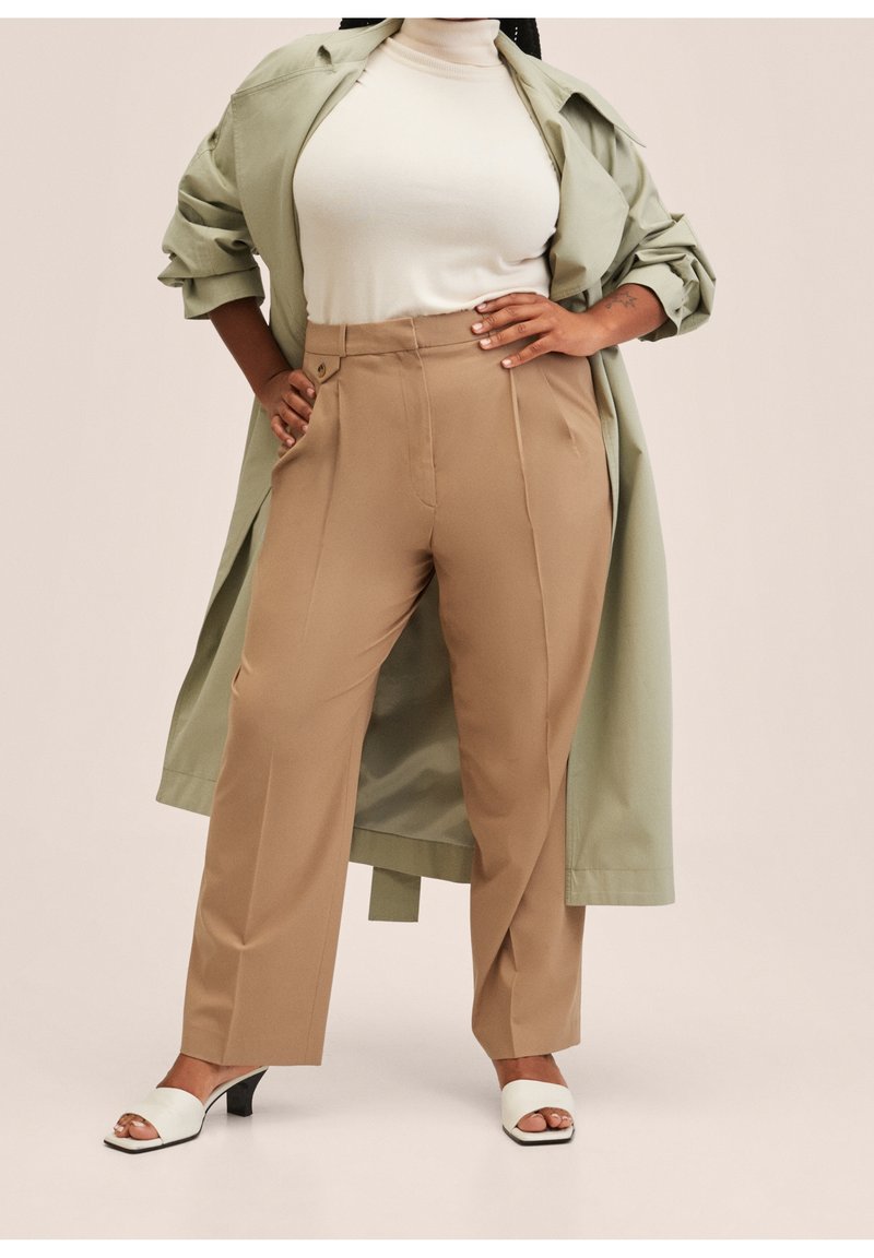 Pantalons beiges ajustés avec des plis à l'avant, un haut crème ajusté assorti et une veste oversize vert clair. Sandales à talons ouverts blanches.