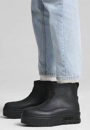 Person iført lyseblå jeans og sorte Puma ankellange regnstøvler med tykke såler og hvide sokker, der er synlige over støvlerne.