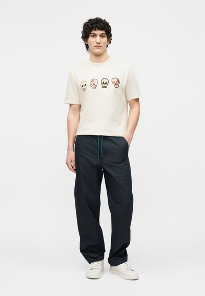 Νεαρός άνδρας με κατσαρά μαλλιά που φορά λευκό μπλουζάκι με τέσσερα πολύχρωμα κρανία, μαύρο παντελόνι με μπλε κορδόνι και λευκά αθλητικά παπούτσια.