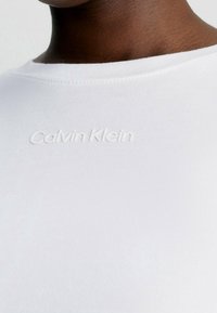 White crewneck t-shirt made of smooth cotton fabric featuring a raised Calvin Klein logo in the center. Seam detailing is visible at the collar.