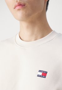 Felpa crema con colletto a coste. Presenta un piccolo logo ricamato "TOMMY JEANS" blu, rosso e bianco sul lato sinistro del petto.