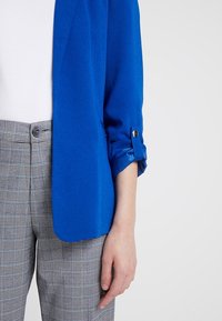 Niebieska marynarka z podwiniętymi rękawami, wykonana z gładkiej tkaniny, zestawiona z szarymi spodniami w kratę, posiadającymi dwa guziki i dopasowany krój.