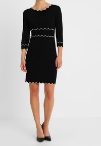 Svart stickad klänning med trekvartsärmar, vågig halsringning och nederkant, vita zigzagdetaljer vid midjan och ärmkanterna.