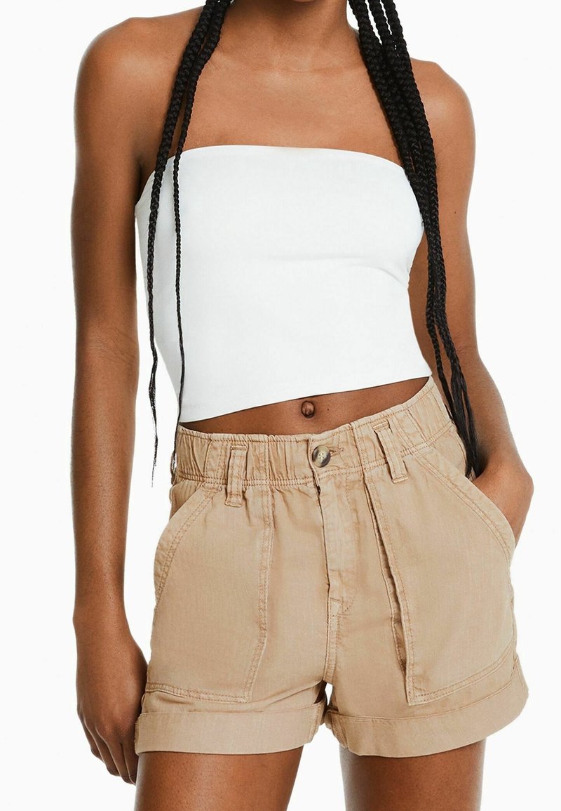 Valkoinen olkaimeton toppi, yhdistettynä vaaleanbeigeihin shortseihin, joissa on napin sulkeminen, taitettavat hihansuut ja kaksi etutaskua.