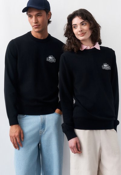 Homme portant un pull Lacoste noir, jean bleu et casquette bleu marine, mains dans les poches ; femme portant un pull Lacoste noir, col rose, pantalon beige, souriante.