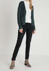Tyrkysový pletený cardigan s dlouhými rukávy, bílý top s vysokým límcem, černé slim kalhoty a vzorované hnědé kotníkové boty s blokovým podpatkem.