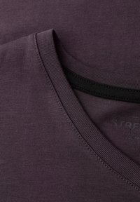 Camiseta morada oscura con cuello redondo, hecha de material suave. Presenta un acabado de costura limpia y una etiqueta discreta con la marca.