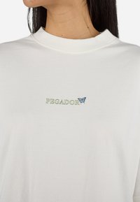 Camiseta de algodón blanca con cuello redondo, que presenta el texto verde "PEGADOR" y una mariposa azul bordada en el pecho.