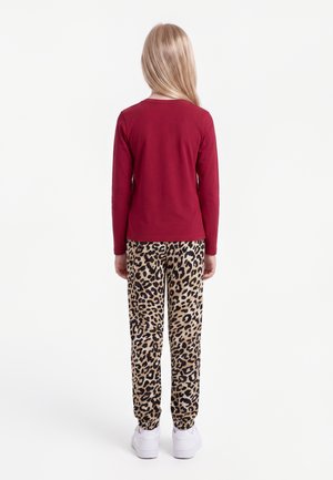 Enfant portant un t-shirt rouge à manches longues, un pantalon imprimé léopard et des baskets blanches, debout de dos sur un fond blanc uni.