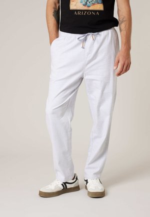 Homme portant un pantalon blanc à cordon, des baskets blanches avec des rayures noires, et un t-shirt graphique noir Arizona, debout les mains détendues.